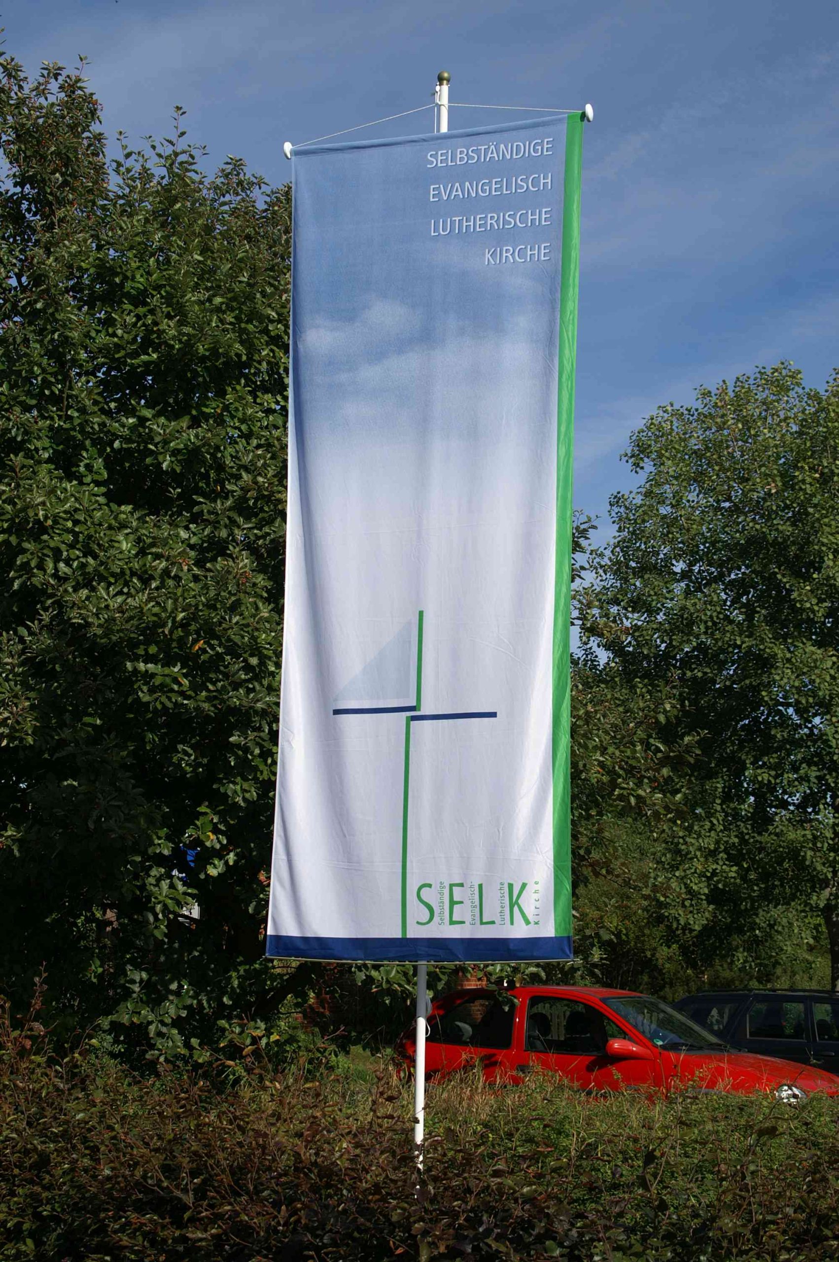 Fahne mit Logo der Selbständigen Evangelisch-Lutherischen Kirche vor dem Parkplatz der Holzkirche